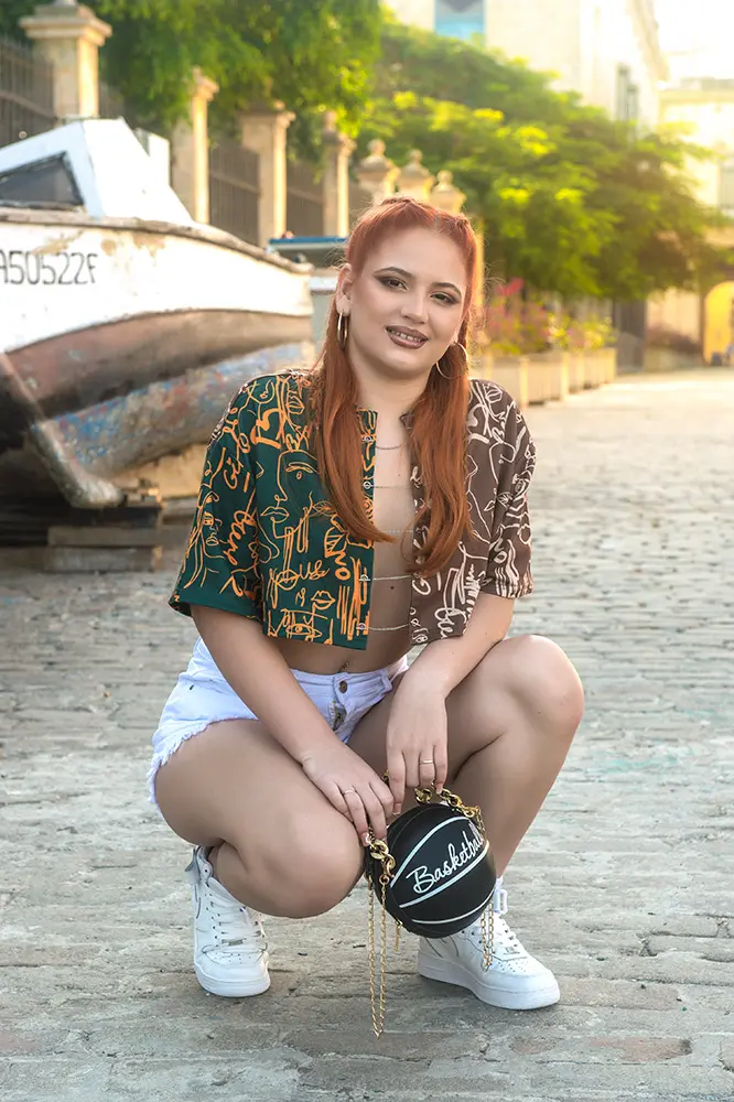 Modelo pelirroja agachada sobre calle de adoquines frente a un bote sobre pilotes en la calle