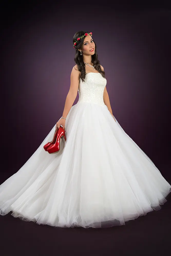 Joven posando con vestido de fantasía blanco y zapatos rojos en la mano